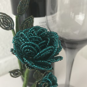 May include: A teal blue beaded rose with green leaves and a stem. The rose is made of small beads and is a delicate and intricate piece of art.