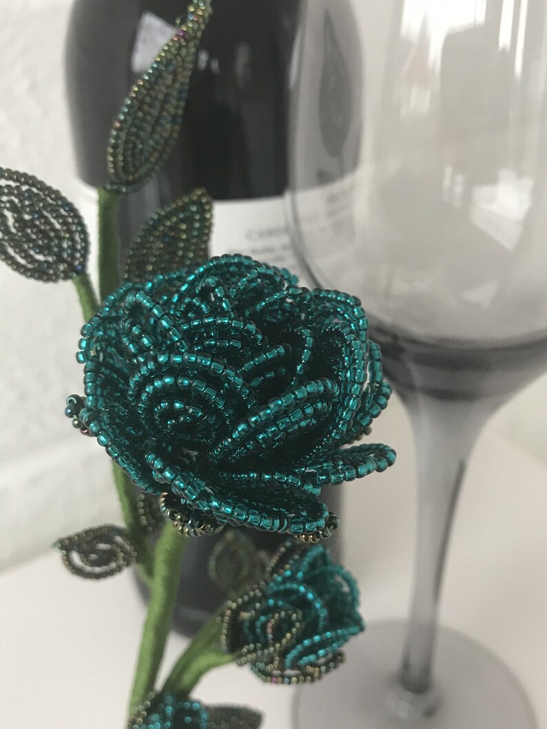 May include: A teal blue beaded rose with green leaves and a stem. The rose is made of small beads and is a delicate and intricate piece of art.