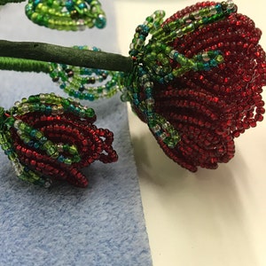 May include: A close-up of a beaded rose made with red, green, and blue beads. The rose is on a blue background.