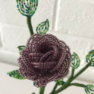 May include: A beaded flower bouquet with a purple rose and green leaves. The rose is made of small, round beads and is arranged in a spiral shape.