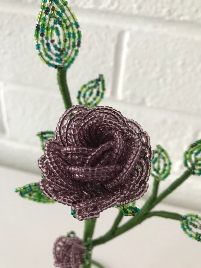 May include: A beaded flower bouquet with a purple rose and green leaves. The rose is made of small, round beads and is arranged in a spiral shape.
