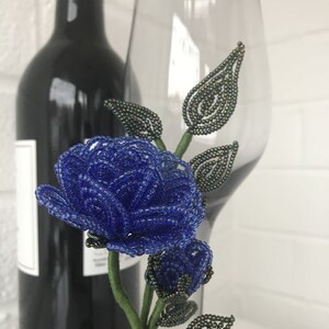 May include: A blue beaded rose with green beaded leaves and stem. The rose is displayed in a clear glass wine glass.