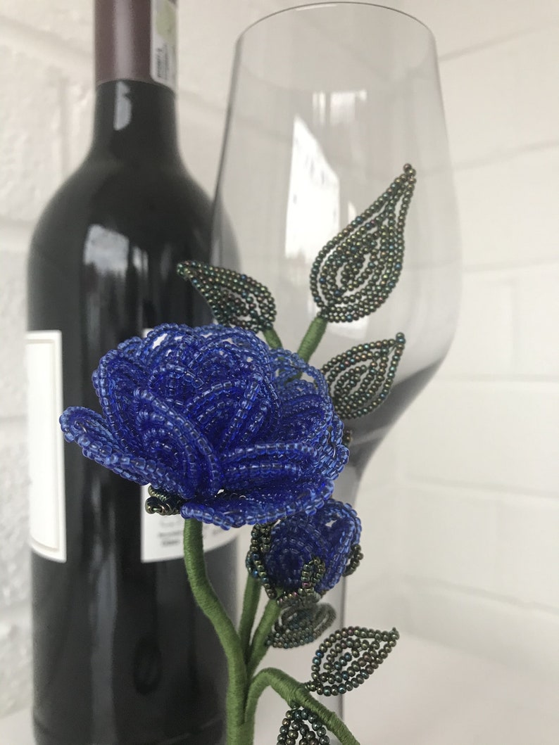 May include: A blue beaded rose with green beaded leaves and stem. The rose is displayed in a clear glass wine glass.