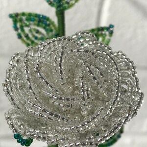May include: A white beaded rose with green leaves. The rose is made of small, clear beads that are strung together to create a realistic-looking flower.
