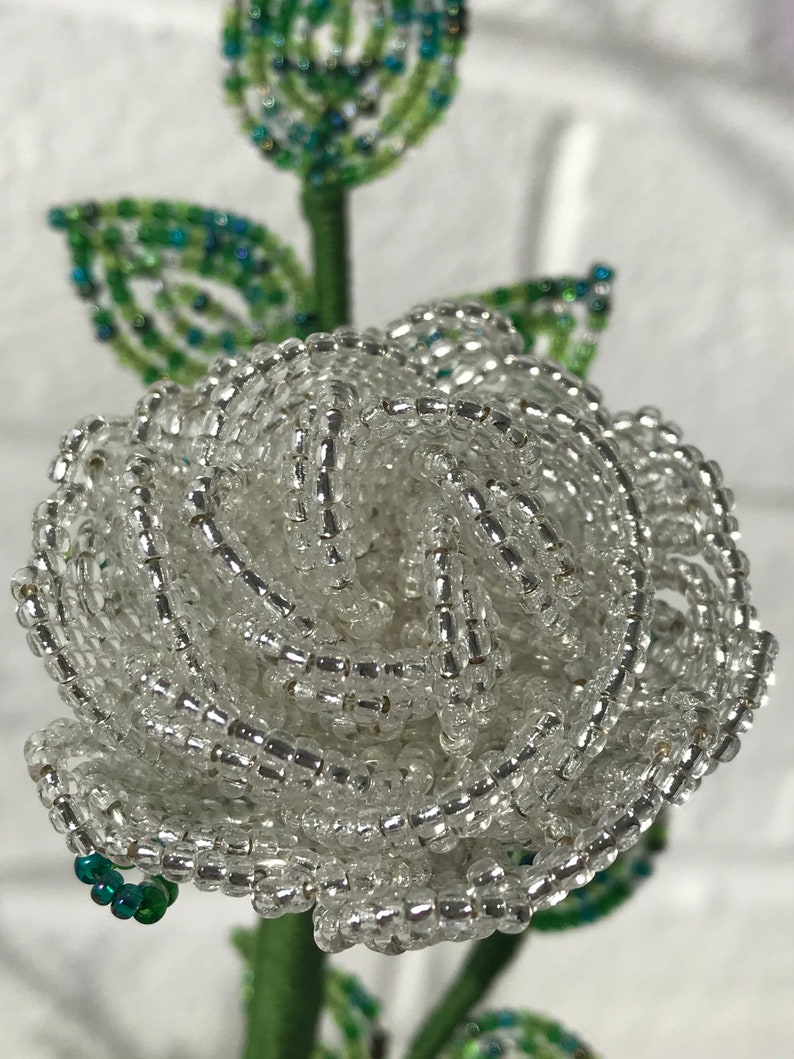 May include: A white beaded rose with green leaves. The rose is made of small, clear beads that are strung together to create a realistic-looking flower.