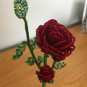 May include: A beaded rose with green leaves and a bud, on a wire stem and base. The rose is red and the leaves are green.