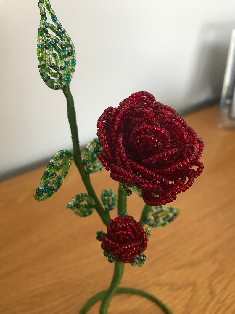 May include: A beaded rose with green leaves and a bud, on a wire stem and base. The rose is red and the leaves are green.