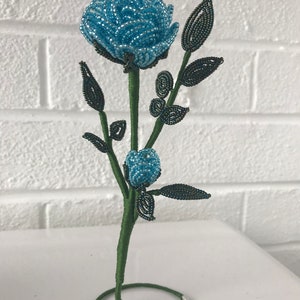 May include: A blue beaded rose with green beaded leaves and stem, standing on a green wire base.