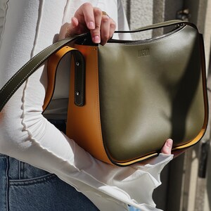 May include: A colour-blocked handbag with olive green, mustard yellow, and black panels. The bag has a long strap and the word "LIMPONI" is embossed on the front. The bag is held by a person wearing a white top and blue jeans.
