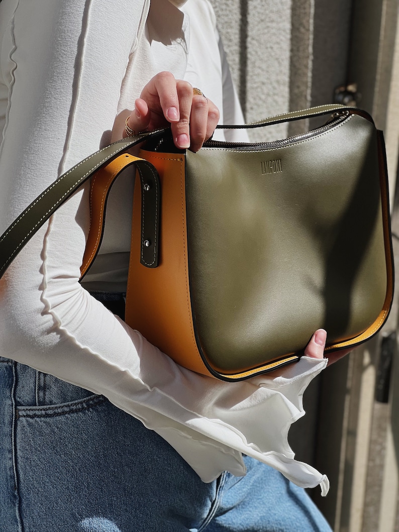 May include: A colour-blocked handbag with olive green, mustard yellow, and black panels. The bag has a long strap and the word "LIMPONI" is embossed on the front. The bag is held by a person wearing a white top and blue jeans.
