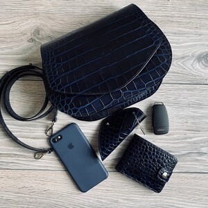 May include: A navy blue leather crossbody bag with a crocodile texture, a matching phone case, wallet, key holder, and car key. The bag has a long strap and silver hardware. The items are arranged on a light wood surface.