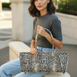 May include: A rectangular handbag featuring a detailed snakeskin pattern in shades of gray, black, and cream. The bag has a structured silhouette, a top flap closure, and two thin, matching snakeskin-patterned handles attached to the sides.