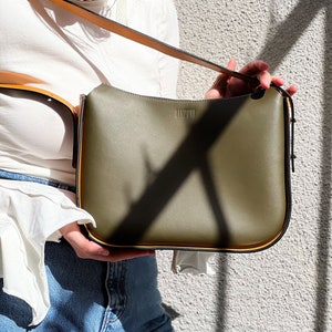 May include: An olive green handbag with a long, adjustable strap. The bag has a rectangular shape with rounded corners and a gold trim. The word "LAMPONI" is printed on the front of the bag. The bag is held against a textured grey wall.