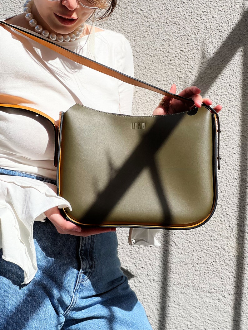 May include: An olive green handbag with a long, adjustable strap. The bag has a rectangular shape with rounded corners and a gold trim. The word "LAMPONI" is printed on the front of the bag. The bag is held against a textured grey wall.