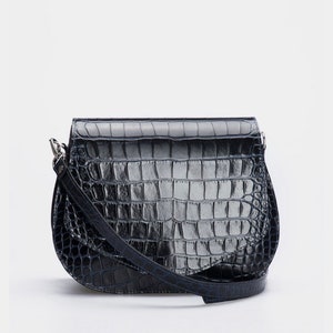May include: A black, faux-crocodile leather crossbody bag with a curved bottom and a flap closure. The bag has a long, adjustable strap and silver-toned hardware. The bag's textured surface gives it a stylish look.