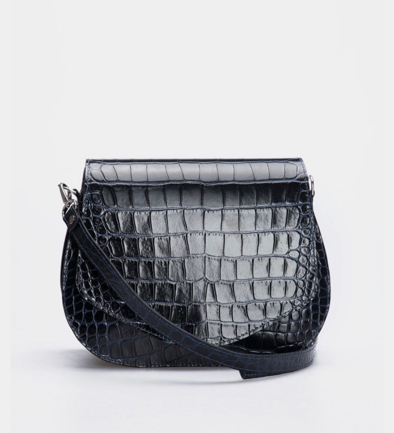 May include: A black, faux-crocodile leather crossbody bag with a curved bottom and a flap closure. The bag has a long, adjustable strap and silver-toned hardware. The bag's textured surface gives it a stylish look.