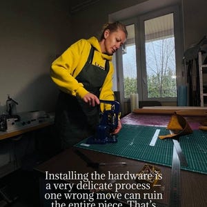 May include: A person in a yellow hoodie and black apron uses a blue press to install hardware on a leather project. The text in the image reads: "Installing the hardware is a very delicate process..."