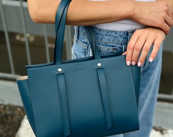 May include: A deep teal blue structured handbag featuring two thin top handles, two vertical decorative leather strips on the front, and silver-toned metal hardware. The brand name LAMESKI is embossed in the center top of the bag exterior.