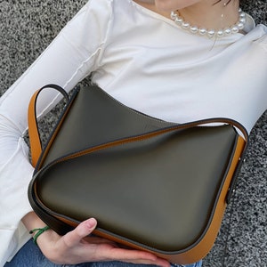 May include: A dark olive green shoulder bag with tan trim and strap. The bag has a curved design. The bag is held by a person wearing a white top and blue jeans. The bag appears to be made of leather. The bag's strap is designed to be worn over the shoulder.
