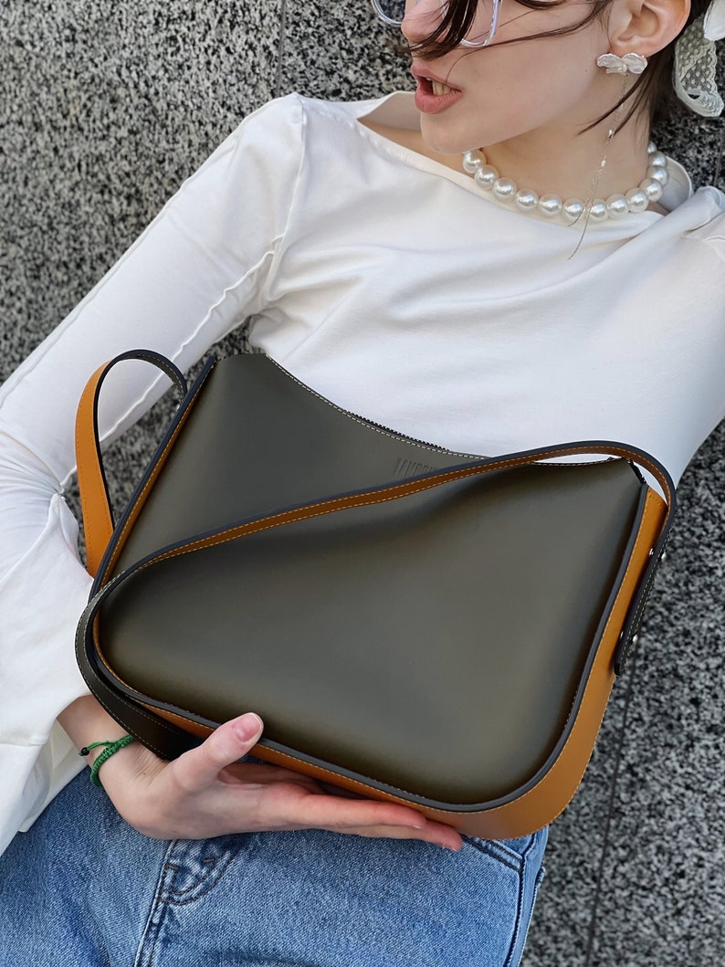 May include: A dark olive green shoulder bag with tan trim and strap. The bag has a curved design. The bag is held by a person wearing a white top and blue jeans. The bag appears to be made of leather. The bag's strap is designed to be worn over the shoulder.