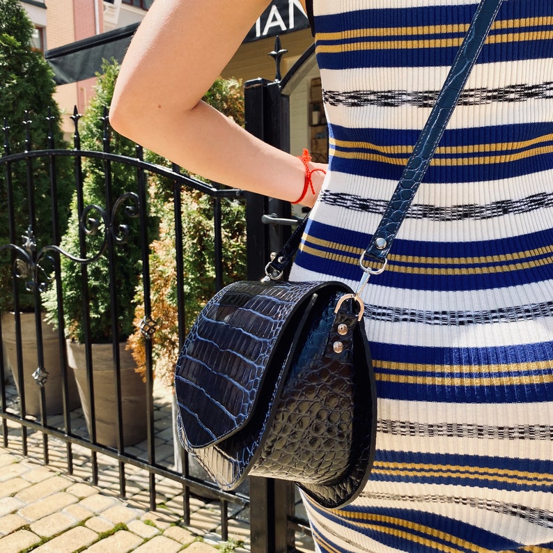 May include: A black, crocodile-textured crossbody bag with a flap closure and a long, adjustable strap. The bag has silver-toned hardware and a curved shape. The strap is attached to the bag with silver rings. The bag is worn over a striped dress.