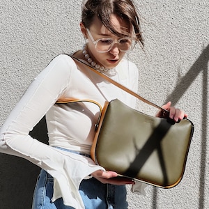 May include: An olive green shoulder bag with tan trim and strap. The bag has a rectangular shape and a minimalist design. The background is a textured grey wall, with sunlight casting shadows.