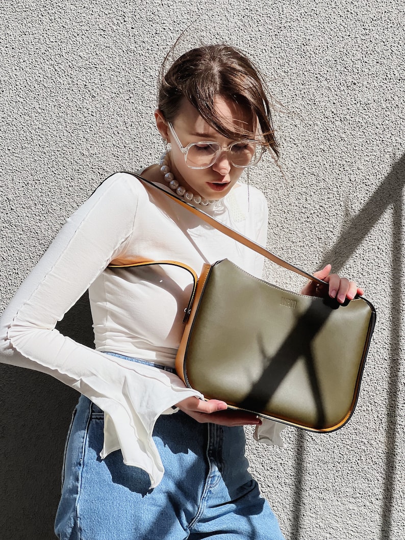 May include: An olive green shoulder bag with tan trim and strap. The bag has a rectangular shape and a minimalist design. The background is a textured grey wall, with sunlight casting shadows.