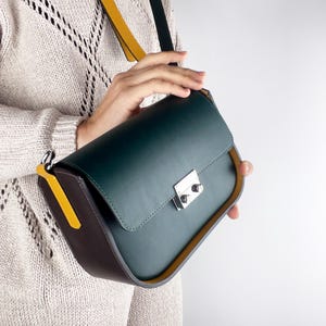 May include: A dark green leather crossbody bag with a silver clasp. The bag has a brown side panel and a yellow strap. The bag is held by a person wearing a light-colored sweater.