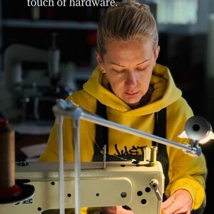May include: A person in a yellow hoodie using a sewing machine. The image includes the text: "I handcraft each product from start to finish - from the first sketch to the final touch of hardware."