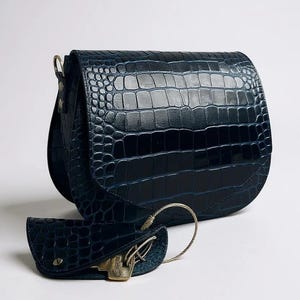 May include: A navy blue, crocodile-textured leather shoulder bag with a matching key holder. The bag has a curved flap and a gold-toned metal clasp. The key holder is attached with a silver-colored metal ring.