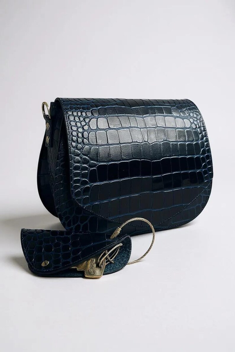 May include: A navy blue, crocodile-textured leather shoulder bag with a matching key holder. The bag has a curved flap and a gold-toned metal clasp. The key holder is attached with a silver-colored metal ring.