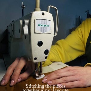 May include: A close-up of a cream-colored ZOJE sewing machine stitching brown leather. The text reads: "Stitching the pieces together is my favorite part of the process - that's when each detail finally comes to life."