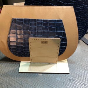 May include: A tan leather bag with a dark blue crocodile-textured pocket and a wooden base. The bag's components are arranged on a wooden surface, with the text "LAMPONI" visible on the wooden base.