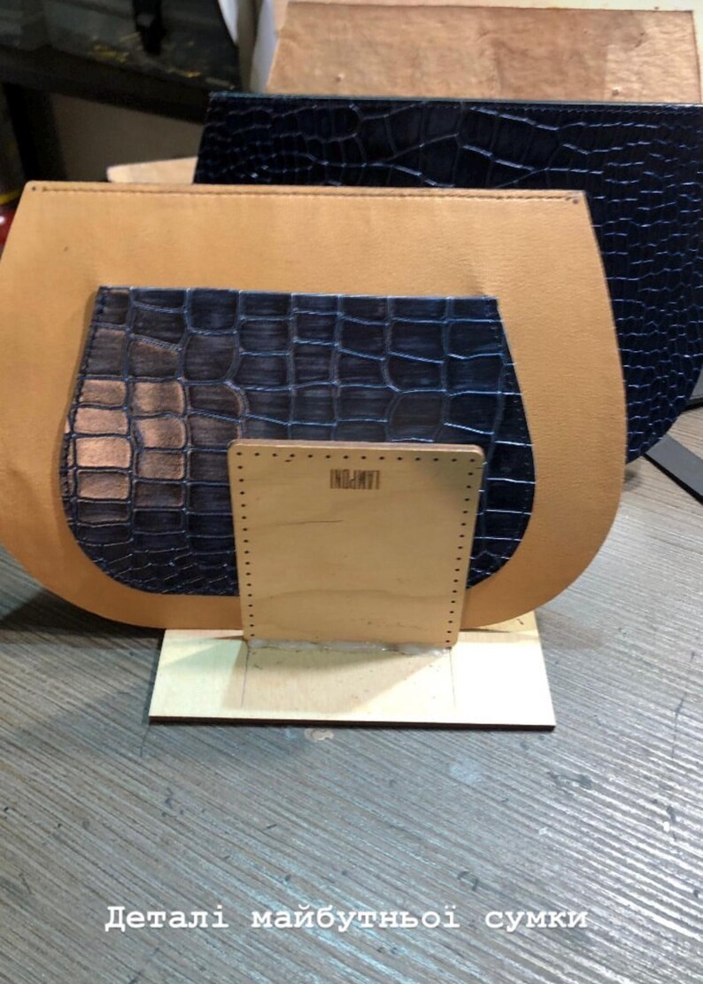 May include: A tan leather bag with a dark blue crocodile-textured pocket and a wooden base. The bag's components are arranged on a wooden surface, with the text "LAMPONI" visible on the wooden base.