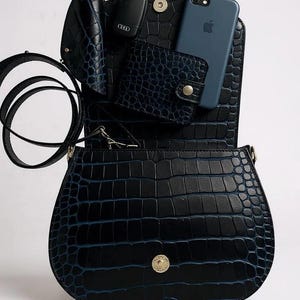 May include: Black faux leather handbag with a crocodile skin texture. The bag is open, revealing a blue phone, wallet, and key holder. The bag has a long strap and a gold-colored clasp.