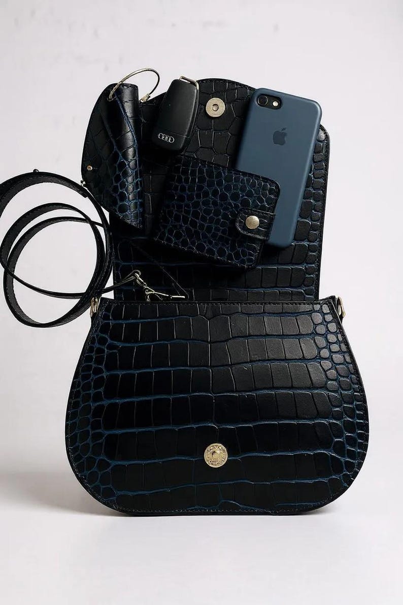 May include: Black faux leather handbag with a crocodile skin texture. The bag is open, revealing a blue phone, wallet, and key holder. The bag has a long strap and a gold-colored clasp.