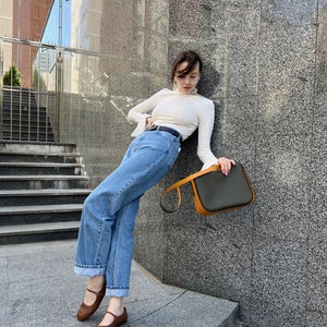 May include: A woman's ensemble comprising a white long-sleeved top, blue jeans, and brown Mary Jane flats. A black belt and a dark green and orange handbag complete the look. The woman is leaning against a grey stone wall.