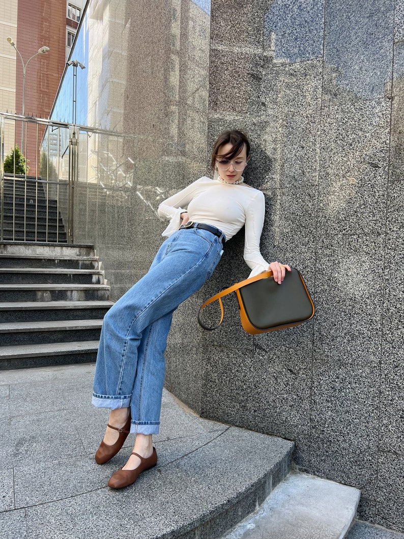 May include: A woman's ensemble comprising a white long-sleeved top, blue jeans, and brown Mary Jane flats. A black belt and a dark green and orange handbag complete the look. The woman is leaning against a grey stone wall.
