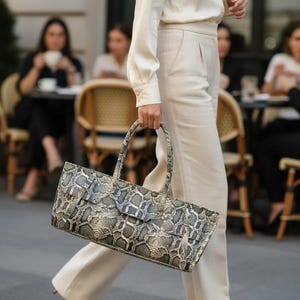May include: A rectangular handbag featuring a grey, black, and white snakeskin pattern. The bag has two rounded top handles and a flap closure with a strap detail across the front. The material has a textured finish with a distinct scale design.