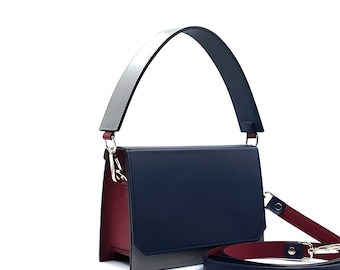 May include: A modern handbag with a rectangular shape. The bag features a navy blue front, a gray bottom section, and a burgundy side. It has a silver and gray handle and a detachable navy blue and burgundy shoulder strap.