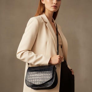 May include: A black, crocodile-embossed leather crossbody bag with a flap closure. The bag has a silver chain strap and is worn over the shoulder. The bag is paired with a cream-colored blazer and black pants.