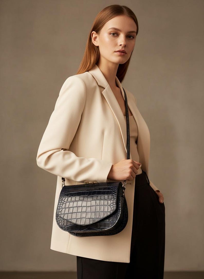May include: A black, crocodile-embossed leather crossbody bag with a flap closure. The bag has a silver chain strap and is worn over the shoulder. The bag is paired with a cream-colored blazer and black pants.