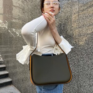 May include: An olive green handbag with a curved top and orange trim. The bag has a long strap. The bag is held by a person wearing a white top and blue jeans.