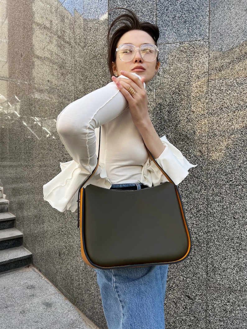 May include: An olive green handbag with a curved top and orange trim. The bag has a long strap. The bag is held by a person wearing a white top and blue jeans.