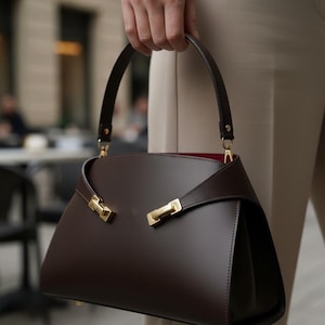 May include: A dark brown structured handbag with a single top handle and gold-tone metal hardware accents. The bag features a smooth finish, a curved top flap design, and a contrasting red interior lining, secured with two gold-colored metal clasps.