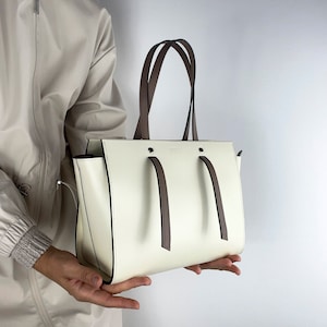 May include: A structured off-white handbag featuring two dark brown top handles and two matching brown decorative straps hanging from the front. The bag has a clean, minimalist design with dark brown edge detailing and a smooth, matte finish.