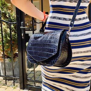 May include: A dark blue, glossy, crocodile-textured handbag with a curved flap and a shoulder strap. The bag is held in front of a person wearing a striped dress with blue, white, and gold stripes. The bag has silver hardware.