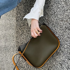May include: Olive green shoulder bag with a contrasting yellow strap and trim. The bag has a rectangular shape with rounded corners and a minimalist design. The strap is long enough to be worn over the shoulder.