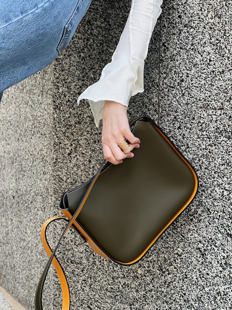 May include: Olive green shoulder bag with a contrasting yellow strap and trim. The bag has a rectangular shape with rounded corners and a minimalist design. The strap is long enough to be worn over the shoulder.