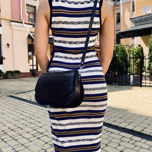 May include: A black, textured leather crossbody bag with a flap closure and a silver-tone chain strap. The bag is worn over a striped dress with white, navy, and gold horizontal stripes. The dress has a sleeveless design.